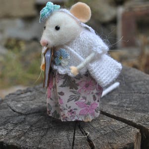 May include: A white felt mouse doll wearing a white knitted cardigan and a floral skirt. The mouse is holding a small book in its paws and has a blue flower on its head.
