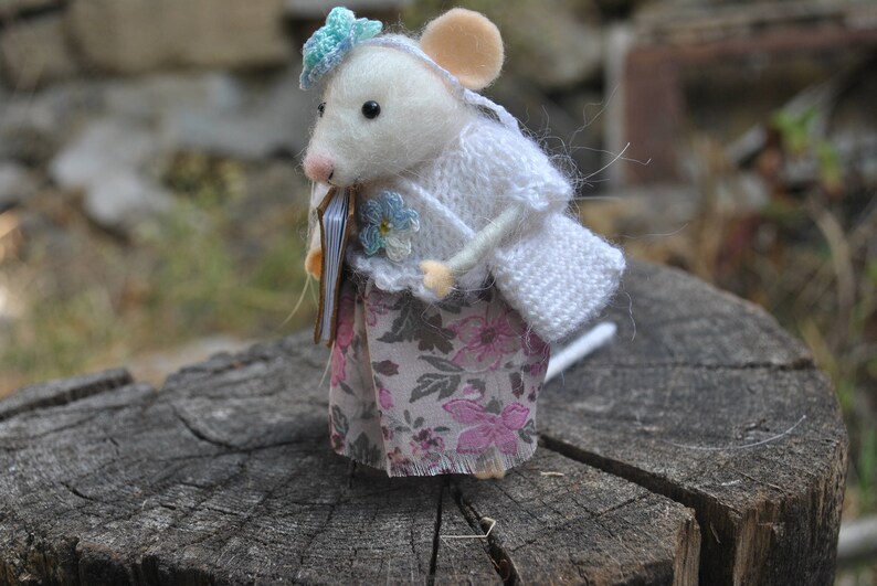 May include: A white felt mouse doll wearing a white knitted cardigan and a floral skirt. The mouse is holding a small book in its paws and has a blue flower on its head.