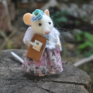 May include: A white felt mouse wearing a blue flower headband and a white knitted sweater holds a brown book with a butterfly design. The mouse is wearing a pink and white floral skirt.