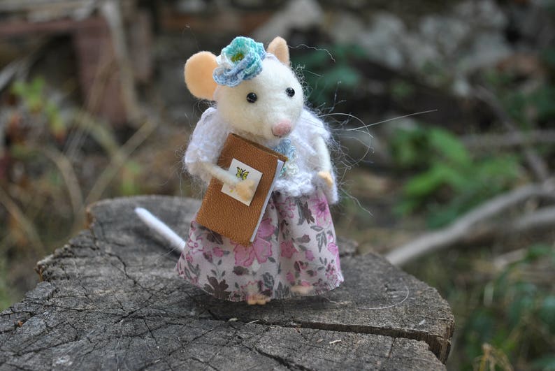 May include: A white felt mouse wearing a blue flower headband and a white knitted sweater holds a brown book with a butterfly design. The mouse is wearing a pink and white floral skirt.