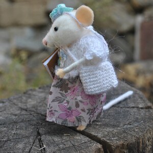 May include: A white felt mouse wearing a white crocheted cardigan and a floral skirt, holding a brown book. The mouse is standing on a tree stump.