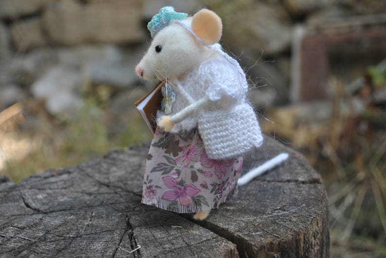 May include: A white felt mouse wearing a white crocheted cardigan and a floral skirt, holding a brown book. The mouse is standing on a tree stump.