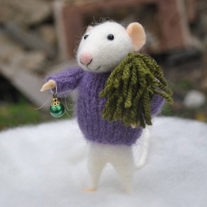May include: A white felt mouse wearing a purple sweater and holding a green ornament. The mouse is standing on a white surface.