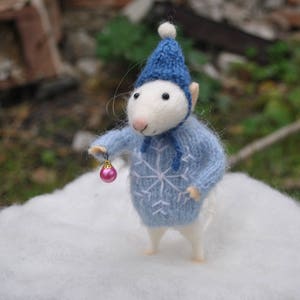 May include: A white felt mouse wearing a blue knitted hat and sweater with a snowflake pattern. The mouse is holding a pink ornament.