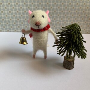 May include: A white felt mouse wearing a red scarf and holding a gold bell, standing next to a small green felt Christmas tree.