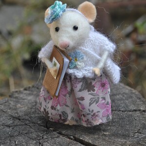 May include: A white felt mouse wearing a blue and white flower hat and a white knitted sweater with a floral skirt. The mouse is holding a brown book.