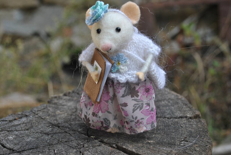 May include: A white felt mouse wearing a blue and white flower hat and a white knitted sweater with a floral skirt. The mouse is holding a brown book.