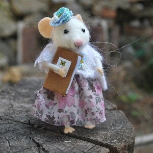 May include: A white felt mouse wearing a blue and white crocheted hat and a pink floral dress. The mouse is holding a brown book with a yellow butterfly on the cover.