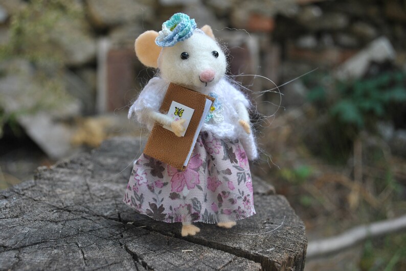 May include: A white felt mouse wearing a blue and white crocheted hat and a pink floral dress. The mouse is holding a brown book with a yellow butterfly on the cover.