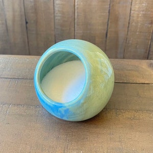 Op de afbeelding: Een bolvormige keramische zoutpot met een grote opening. De buitenkant is geglazuurd in groen- en blauwtinten, met een gladde, glanzende afwerking. De binnenkant is gevuld met wit zout. Een decoratief keukenartikel.