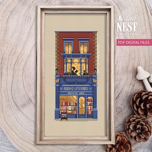 Op de afbeelding: Kruissteekpatroon van een boekwinkel en café, omlijst in licht hout. Het gebouw heeft een rode bakstenen gevel, blauwe accenten en de tekst "BOOKS UPSTAIRS". De ramen zijn verlicht en er zijn katten silhouetten. Het bord zegt "OPEN LATE".