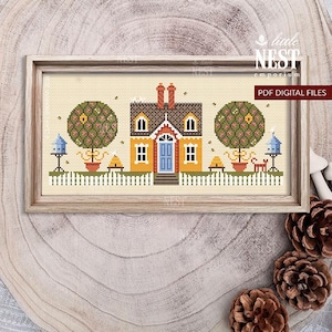 May include: Cross-stitch design of a house with a blue door, yellow walls, and a brown roof, framed in light wood. The scene includes trees, birdhouses, and a picket fence. The text "PDF DIGITAL FILES" is visible.