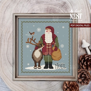 May include: A cross-stitch pattern featuring a bearded Santa Claus in a red and green coat standing next to a reindeer on a light blue background with white snowflakes. The design includes a decorative border and the text NOËL at the bottom.