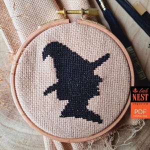 May include: A cross-stitch embroidery hoop featuring a black silhouette of a witch. The witch is wearing a pointed hat. The embroidery is set in a wooden hoop. The text "little NEST emporium PDF DIGITAL FILE" is in the lower right corner.