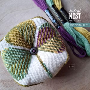 May include: A square, handmade pincushion with a button in the centre, featuring a geometric design in green, purple, and yellow thread on a white background. The image includes spools of embroidery floss and the text "little NEST emporium PDF DIGITAL FILES."