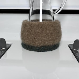May include: A clear glass pitcher featuring a thick, textured, light brown felted wool sleeve around its base, with a darker charcoal gray felted wool band at the very bottom edge, providing a contrasting layered appearance to the vessel.