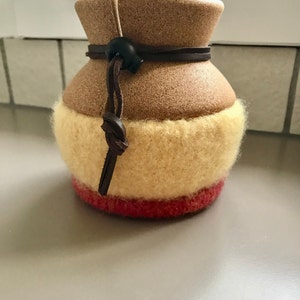 May include: A brown ceramic coffee pot with a beige and red knitted cozy. The cozy has a brown leather strap with a black buckle.