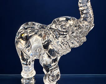 Könnte beinhalten: Eine Elefantenfigur aus klarem Glas im Profil mit erhobenem Rüssel. Die Skulptur weist detaillierte, geformte Texturen auf, die die Haut, Ohren und Stoßzähne des Tieres darstellen, mit einer glatten, reflektierenden Oberfläche am gesamten Körper.