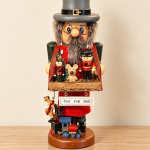 May include: A wooden nutcracker figure wearing a gray top hat and glasses, holding a tray with two small soldier figurines and a bear. A sign hanging below reads TOYS FOR SALE, with a rocking horse and a toy train resting on the base.