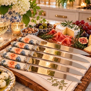 May include: A set of five steak knives with white ceramic handles featuring a floral pattern of orange and blue flowers. Each knife has a serrated stainless steel blade with a visible logo engraving, arranged horizontally on a linen cloth.