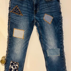 May include: Blue denim jeans with decorative patches in various patterns. Patches include white polka dots on a navy triangle, blue gingham, and a white and blue striped rectangle. The jeans have frayed edges.