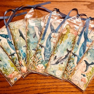 May include: A collection of whale-themed bookmarks. Each bookmark features a watercolor illustration of whales, marine plants, and sea creatures in shades of blue, green, and beige. They are encased in clear plastic with a blue ribbon attached.