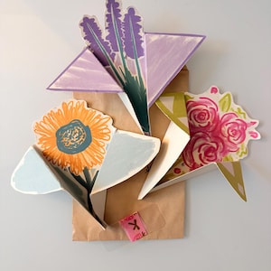 Könnte beinhalten: Eine braune Papiertüte, die mit dreidimensionalen Papierblumen verziert ist. Die Ausschnitte zeigen einen violetten Lavendelzweig, eine gelbe Sonnenblume mit blauem Zentrum und ein Bündel rosa Rosen, die an der Vorderseite befestigt sind.