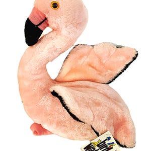 May include: A plush flamingo toy with soft light pink fur, a long curved neck, and a black and pink beak. The bird has orange eyes and black-tipped wings. A white tag attached to the base features the text Bird Watchers and the brand name Dakin.