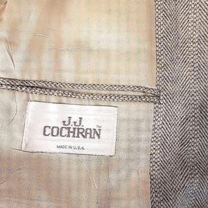 May include: Close-up of the interior of a tweed blazer. The lining is a light beige color, and the label reads "J.J. Cochran Made in U.S.A." The outer fabric is a gray and white herringbone pattern.
