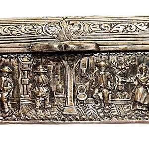 May include: A silver-toned rectangular box with ornate relief carvings. The lid and sides feature detailed scenes of figures in a tavern setting, with decorative floral patterns. The box rests on four small, rounded feet.