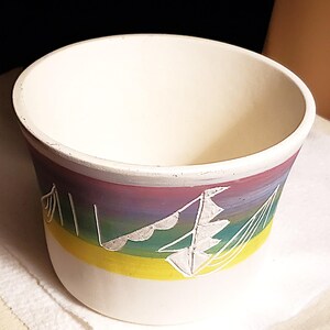 May include: A white ceramic planter with a colorful painted design. The planter features a rainbow gradient of purple, pink, green, and yellow. A white sailboat design is painted on the side. The planter has a wide opening and a rounded base.