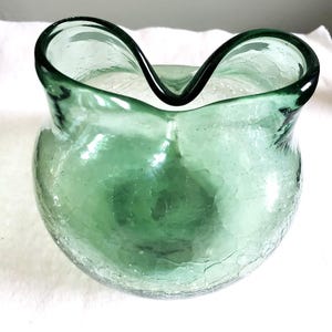 May include: A clear, green glass vase with a unique, wavy rim. The vase has a round body and a textured, crackled appearance. The glass is a soft, sea-green color, and the vase is likely handmade.