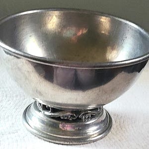 May include: A small, silver-colored bowl with a rounded shape and a decorative base. The bowl has a smooth, reflective surface and sits on a pedestal base with ornate detailing. The bowl is likely made of metal and appears to be vintage.