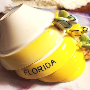 May include: A stack of three ceramic measuring cups with a white top cup and two yellow bottom cups. The middle cup features the word FLORIDA in black text. Each cup has a small, colorful, sculpted bird figurine attached to the handle area.