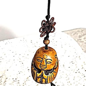 May include: A small, brown, smiling Buddha pendant with black cord and decorative knot. The pendant is a light brown color with dark brown etched details. The cord has two small wooden beads at the bottom.