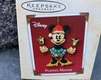 May include: Hallmark Keepsake Ornament featuring Disney's Playful Minnie. The ornament shows Minnie Mouse in a Santa hat and holding bells. The ornament is in a decorative box with the Disney logo.