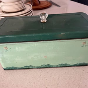 May include: A rectangular metal bread box featuring a dark green lid with a clear glass knob handle. The base is a lighter mint green color, decorated with small floral accents and a scalloped dark green border along the bottom edge.