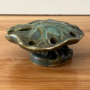 May include: A ceramic flower frog in a leaf shape with a green and brown glaze. The frog has multiple holes for arranging stems and sits on a pedestal base. The piece is on a wooden surface.