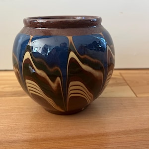 May include: A round ceramic vase with a dark brown rim and base. The body of the vase is a deep blue with a flowing pattern of cream and brown. The vase sits on a wooden surface.