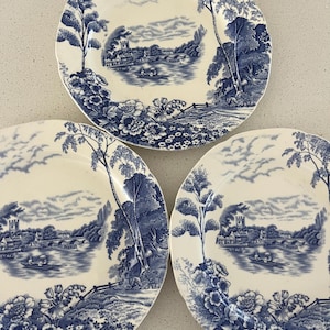 May include: Three round ceramic plates featuring a blue and white transferware pattern. The design depicts a riverside landscape with a stone bridge, a church, and people in a small boat, surrounded by detailed floral borders and tall trees.