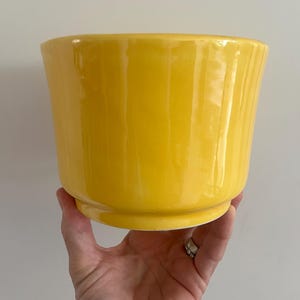 Peut inclure: Un cache-pot en céramique jaune vif avec une finition brillante et une surface légèrement texturée à côtes verticales. Le récipient présente une large ouverture sur le dessus et une base circulaire surélevée, affichant une couleur unie éclatante.