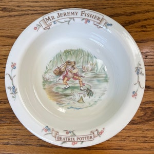 May include: A white ceramic bowl with a blue and green illustration of a frog fishing. The bowl has a brown banner with the text "Mr. Jeremy Fisher" and a smaller banner with the text "Beatrix Potter".