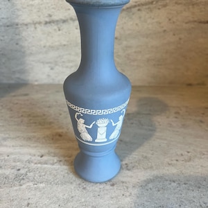 May include: A light blue ceramic vase with a flared rim and base. The vase features a white decorative band with a Greek key pattern and white figures. The vase is approximately 6 inches tall.