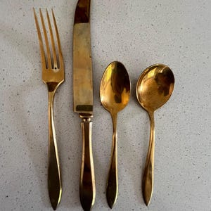 May include: A set of four gold-colored flatware pieces: a fork, a knife, and two spoons. The fork has four tines, and the knife has a rounded blade. The spoons are different sizes. The flatware is arranged on a light gray surface.