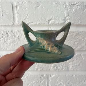 May include: A green ceramic candle holder with a circular base and two curved handles. The candle holder has a textured design on the front and is likely vintage. The color is a mix of green and brown.