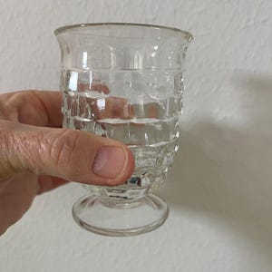 May include: A clear, vintage-style glass with a textured, grid-like pattern on the body. The glass has a rounded top and sits on a small, circular base. The glass is held by a hand, suggesting its size and use.