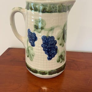 May include: A ceramic pitcher with a cream-colored body, adorned with blue grape clusters and green leaves. The rim and base are trimmed with green, and it has a curved handle. The pitcher has a textured surface.