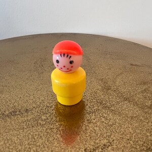 May include: A yellow plastic toy person with a red hat and a pink face. The toy has a simple, smiling face with black eyes and a black line for a mouth.