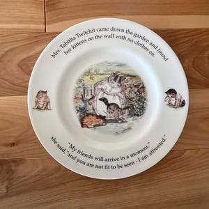 May include: A white ceramic plate with a colorful illustration of a cat and kittens. The plate has a quote around the edge that reads: "Mrs. Tabitha Twitchit came down the garden and found her kittens on the wall with no clothes on. "My friends will arrive in a moment." she said, "and you are not fit to be seen - I am affronted."