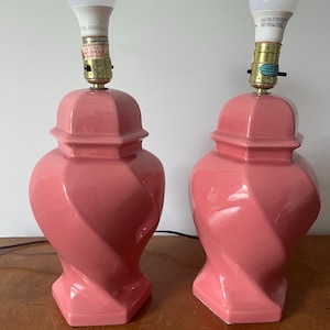 Pode incluir: Duas luminárias de mesa de cerâmica rosa combinando com lâmpadas brancas. As luminárias têm um design torcido exclusivo na base e um formato de urna clássico. As luminárias estão colocadas em uma superfície de madeira.